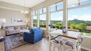 A spacious living room with a large window offering a scenic view, a blue sofa, a white table with chairs, and a decorative rug. - Lake View Condo with 2 K Beds, Golf & Ticket Discounts! (Branson)
