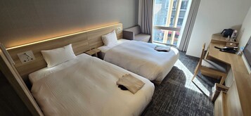 A hotel room with two beds, a desk, a chair, and a window with a view of buildings.