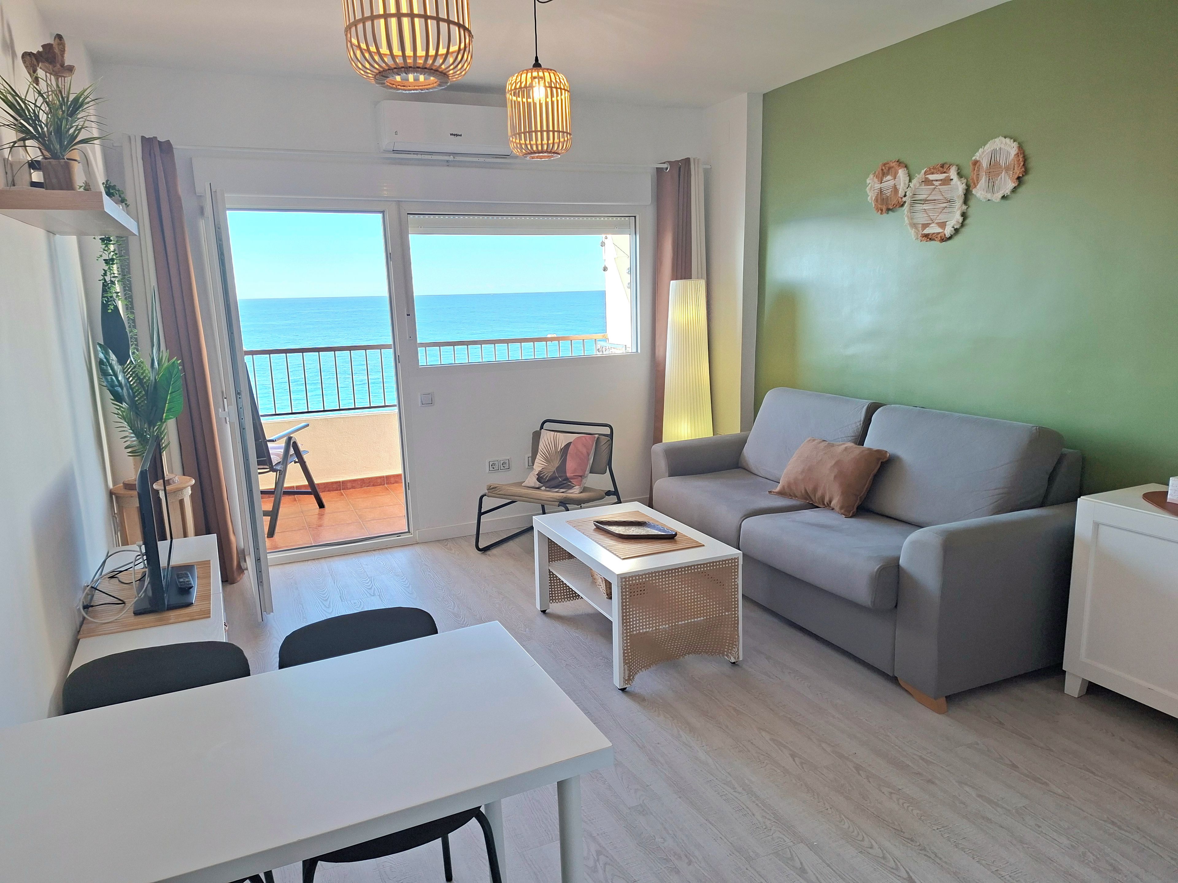 Ein modernes Wohnzimmer mit grauem Sofa, einem weißen Couchtisch und einem Essbereich mit weißem Tisch und Stühlen. Ein Balkon bietet Blick auf das Meer.