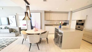 A modern kitchen with a central island, a dining area with chairs, and a TV in the background. - 2-Bedroom Luxury Condo with Balcony (Hollywood)