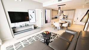 A modern living room with a large flat-screen TV, a sofa, a coffee table with fruit, and a patterned rug. - 2-Bedroom Luxury Condo with Balcony (Hollywood)
