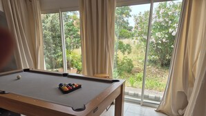 A pool table with balls, a cue stick, and a pool table cover in a room with large windows and curtains. - Petite Maison en Pleine Nature Avec Piscine (Puget-sur-Argens)
