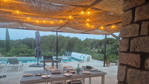 An outdoor dining area with a table set for four, surrounded by chairs, under a canopy with string lights. A pool is visible in the background. - Petite Maison en Pleine Nature Avec Piscine (Puget-sur-Argens)