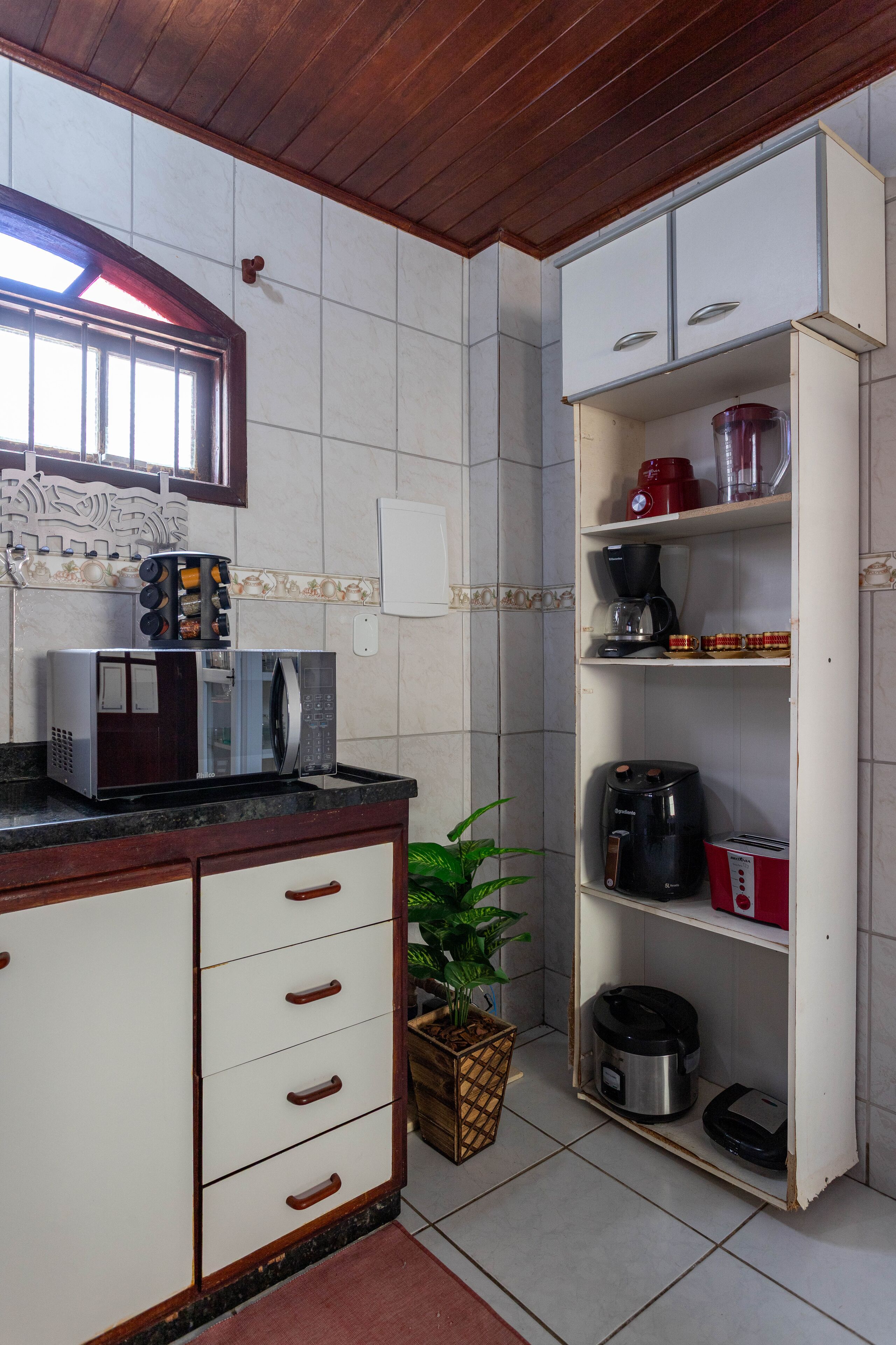 A kitchen with a microwave, coffee maker, and toaster on the counter, a potted plant, and a cabinet with a red appliance on the shelf.