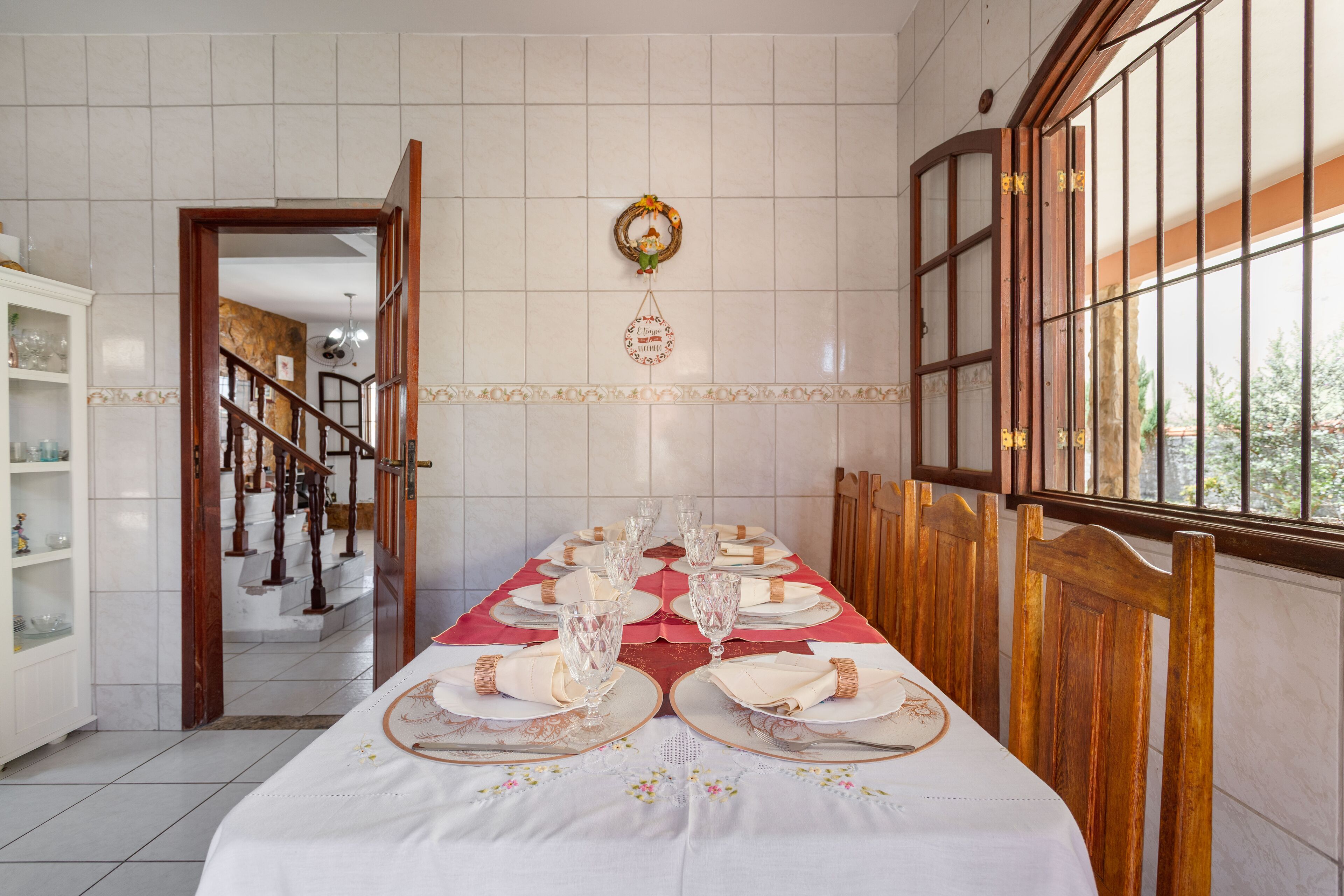 A dining area with a long table set for a meal, a staircase in the background, and a view through a window with bars.