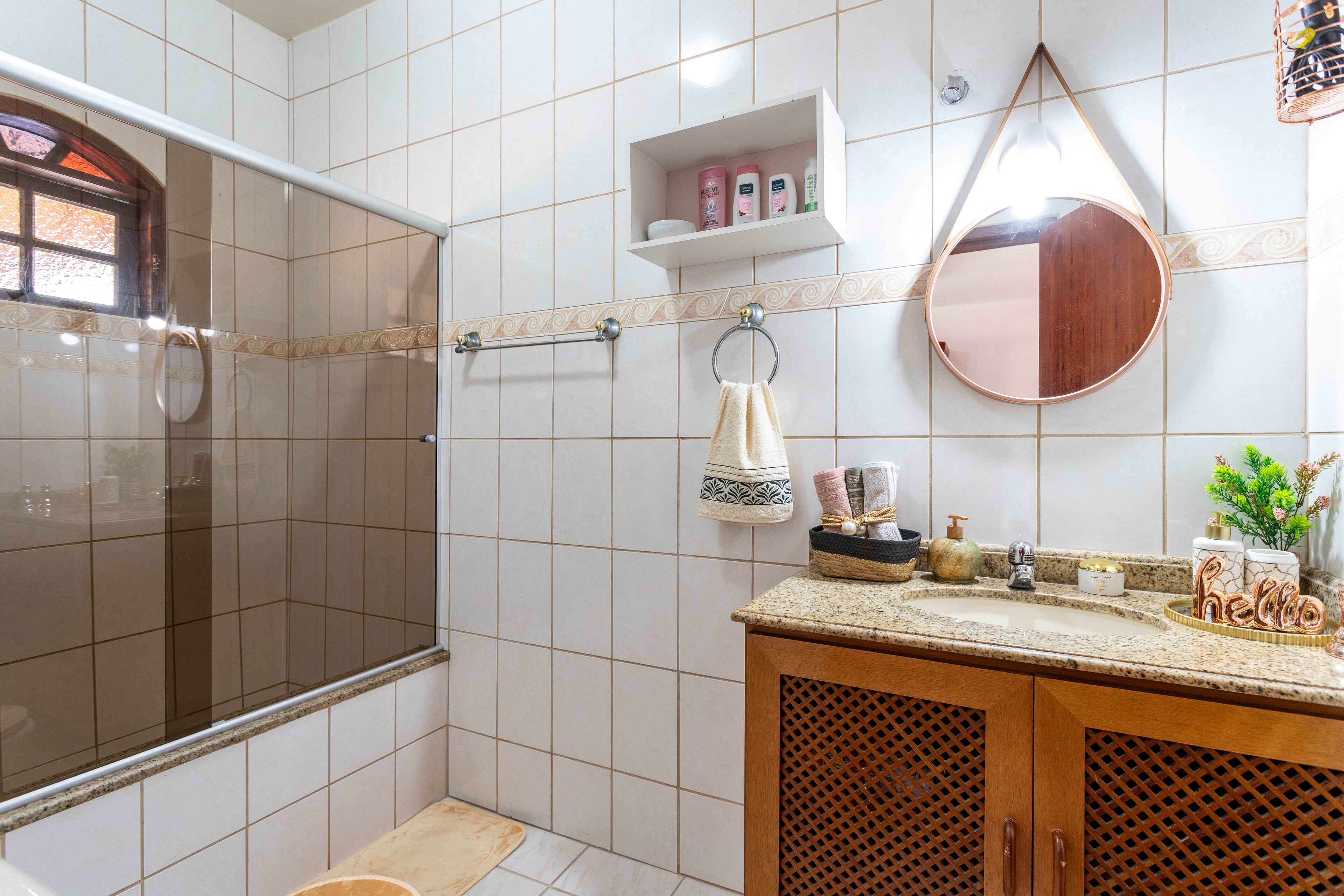 Baño con ducha, lavamanos y espejo. Hay artículos de aseo en el lavamanos y una toalla colgada en la pared.