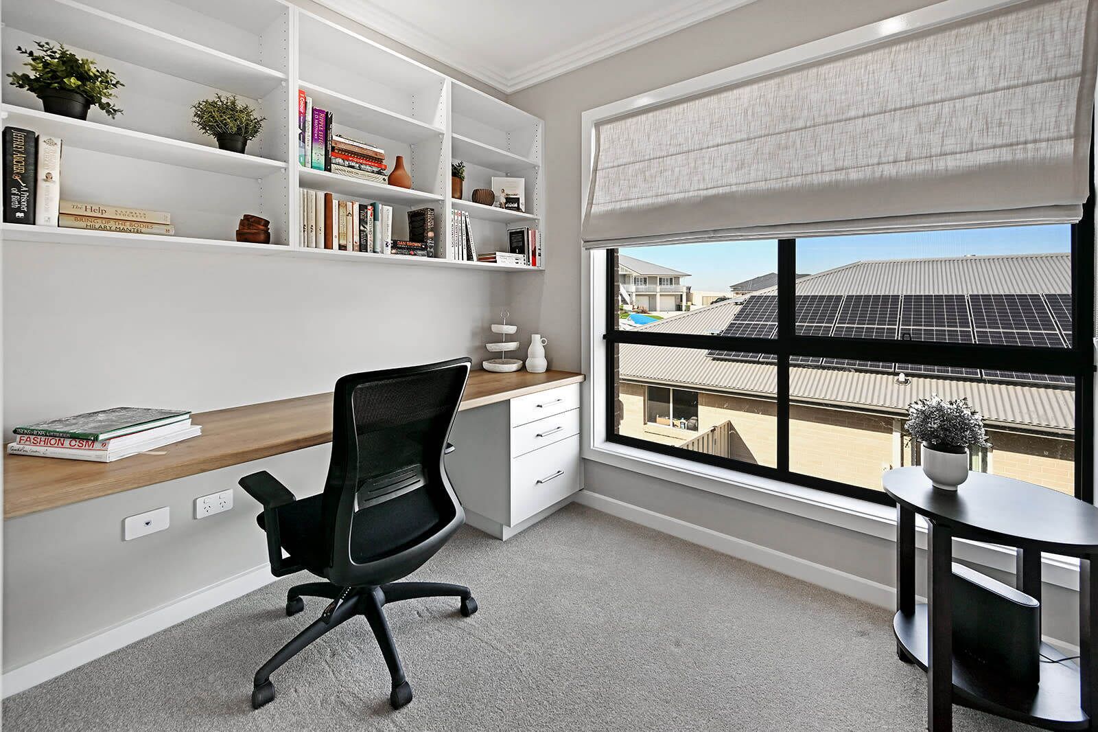 A modern office space with a desk, chair, shelves with books, a window with blinds, and a view of solar panels outside.