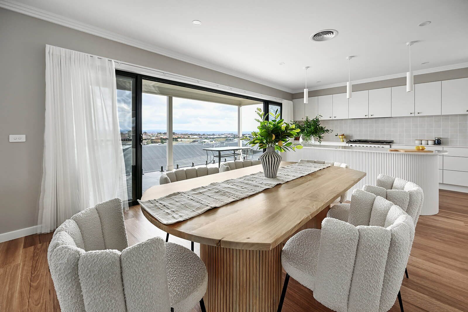 A modern dining area with a wooden table, white chairs, and a view of the city through large windows.