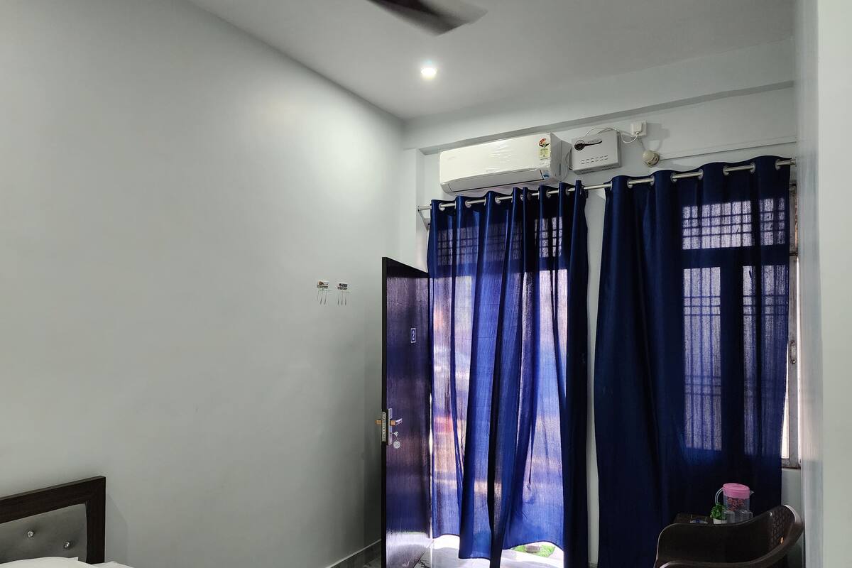 A hotel room with a bed, a window with blue curtains, an air conditioning unit, and a small table with a water bottle.