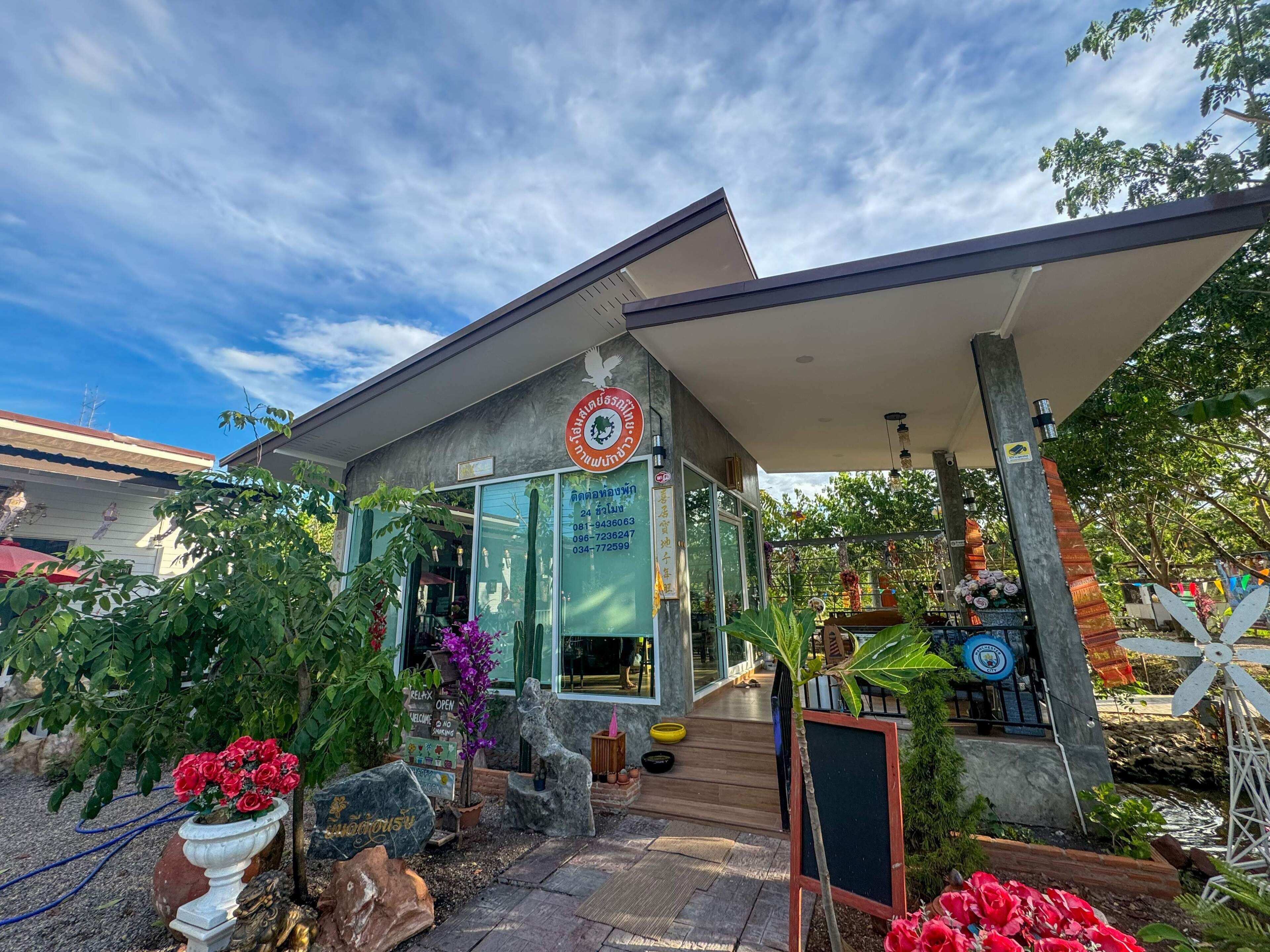 A modern building with a glass facade, a signboard with contact information, and a covered outdoor area with plants and a water feature. - Toraneethai Homestay Amphawa (Amphawa)