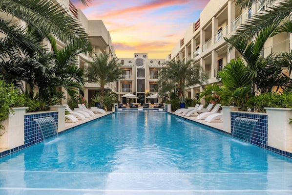 Une piscine avec des chaises longues, des palmiers et un bâtiment doté d’une entrée centrale.