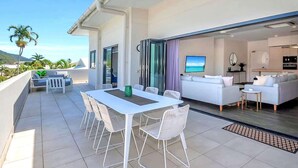 A modern outdoor living space with a dining area, a sofa, and a kitchen area visible through an open sliding door.