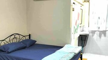 Una cama individual con colchón y almohadas azules, un equipo de aire acondicionado encima y una mesita de noche con lámpara.