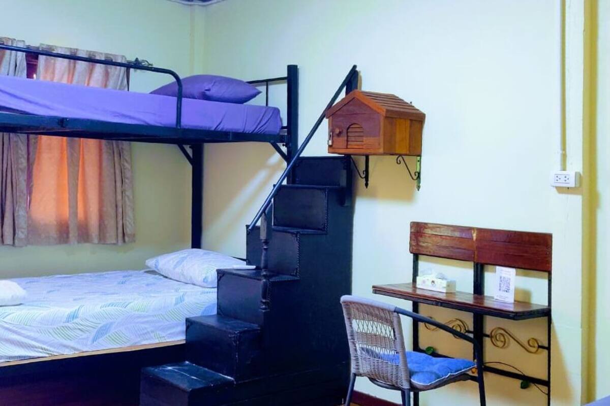 A bunk bed room with a wooden ladder, a small wooden birdhouse, a wicker chair, and a small table with a box and a sign.