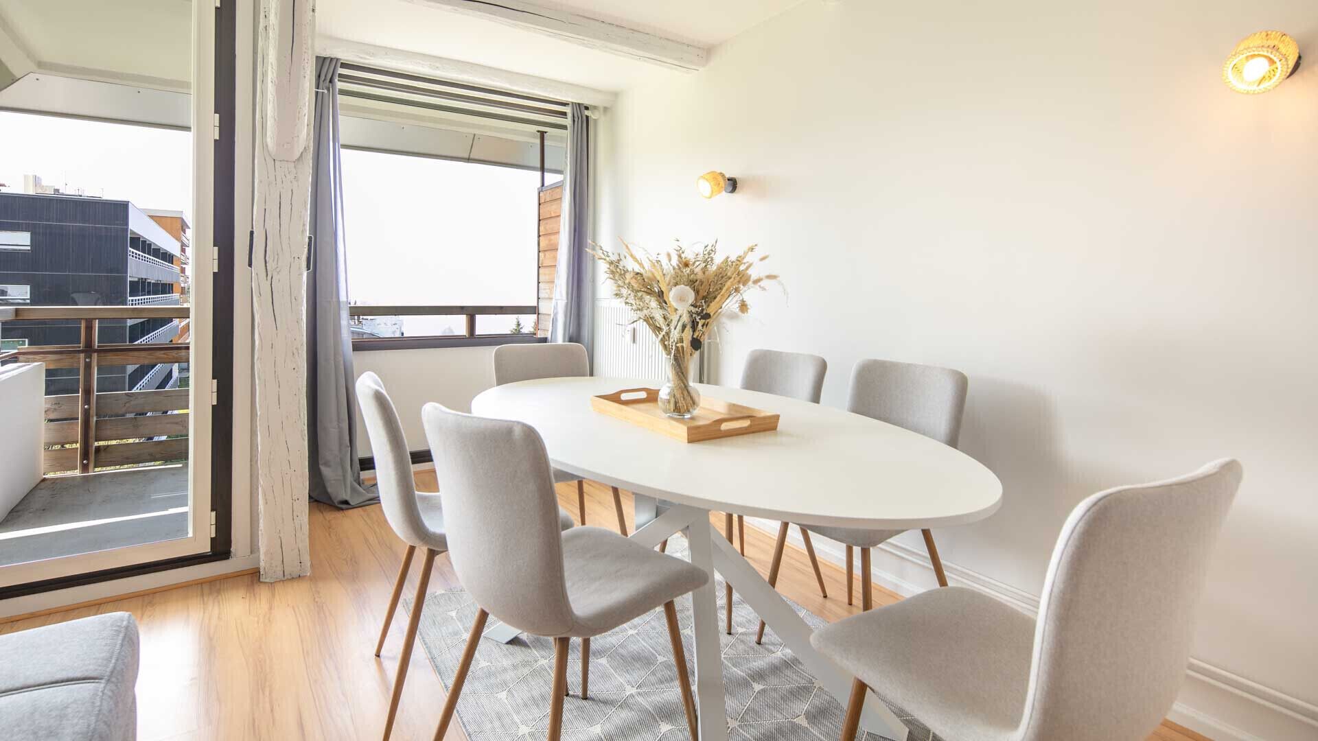 A dining area with a white oval table, six grey chairs, and a vase with dried flowers.