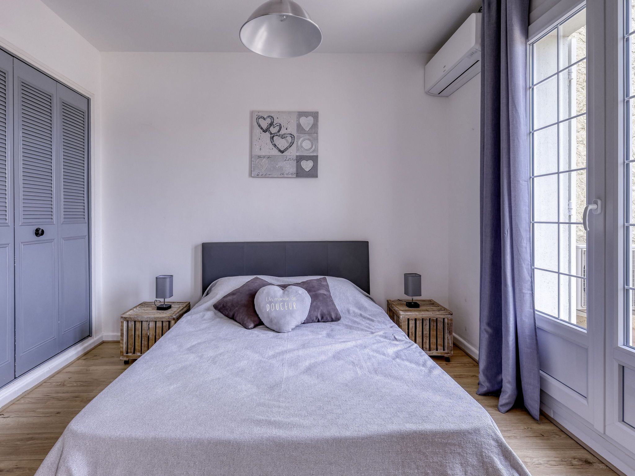 Une chambre à coucher comprenant un lit, deux tables de chevet, un climatiseur fixé au mur et une fenêtre avec des rideaux.
