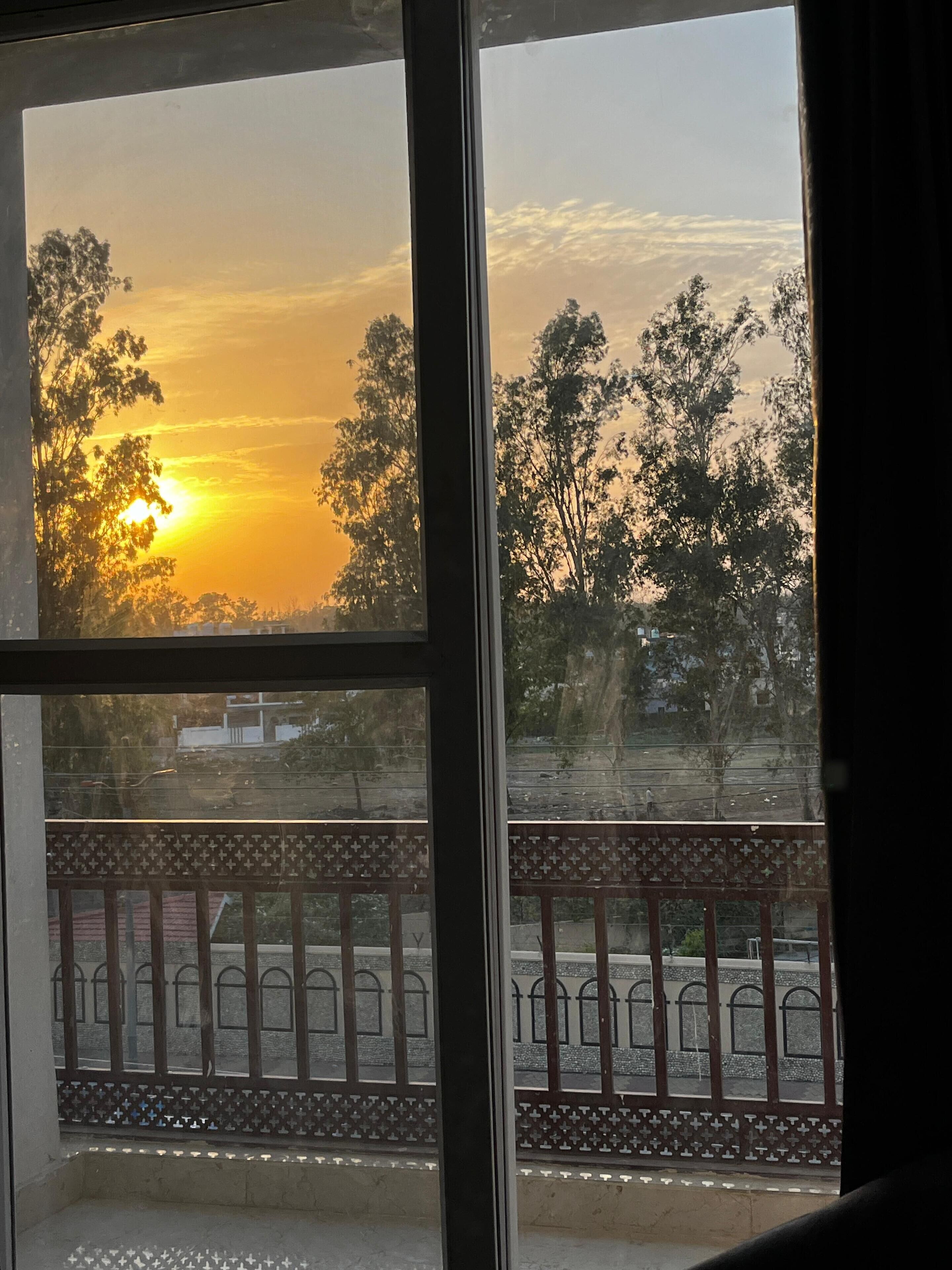 Vista pela janela com varanda, mostrando um pôr do sol e árvores.