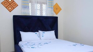Una cama con cabecera oscura, ropa de cama blanca con motivos florales azules y una ventana con cortinas estampadas.