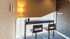 A small, modern room with a glass wall, a table, two chairs, a lamp, and a ceiling exit sign. - Tabist Takamatsu Pearl Hotel (Takamatsu)