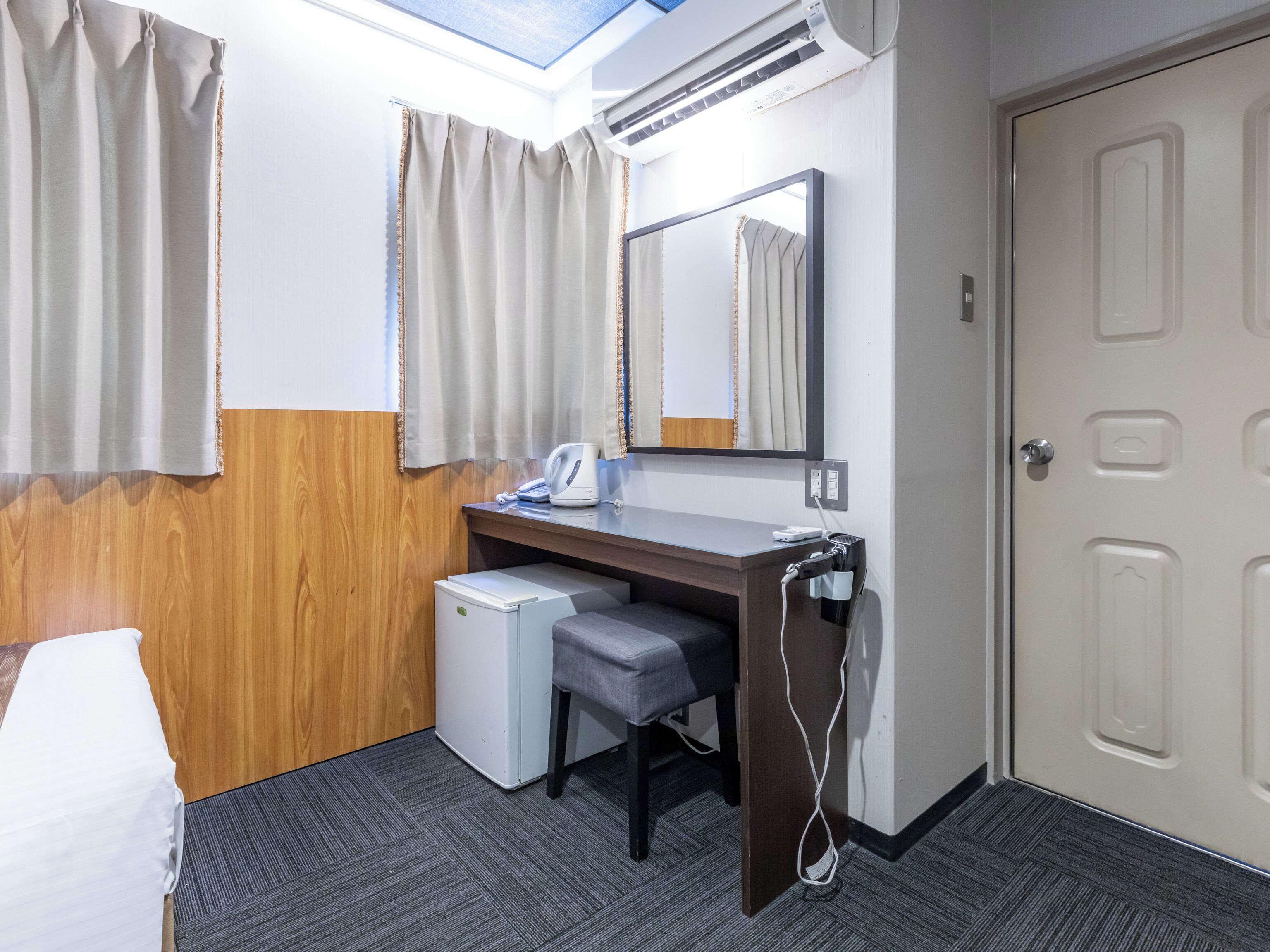 A hotel room with a bed, a desk with a chair, a small refrigerator, a mirror, and a door.