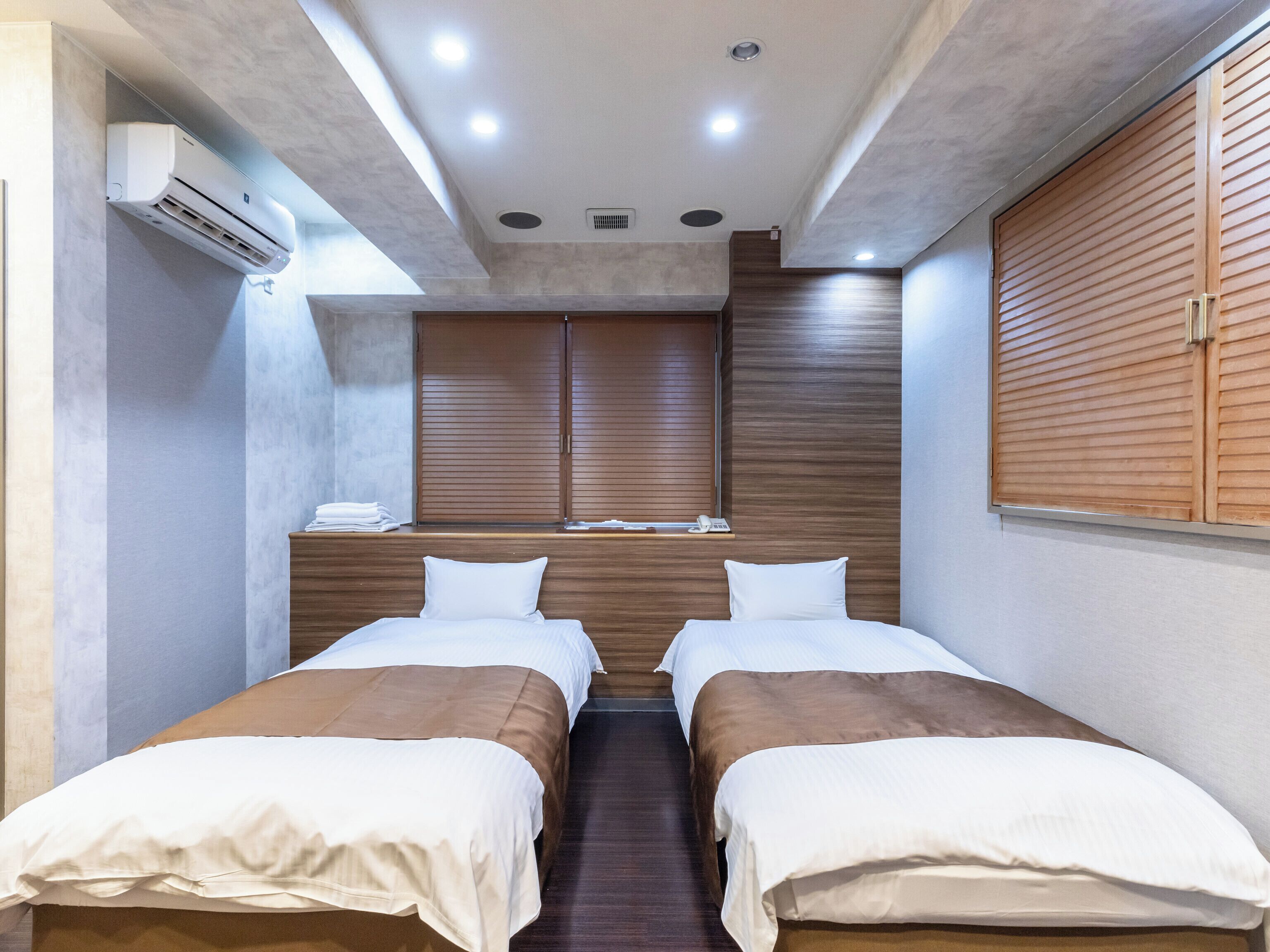 A modern hotel room with two single beds, a wooden wardrobe, a built-in air conditioning unit, and a window with blinds.