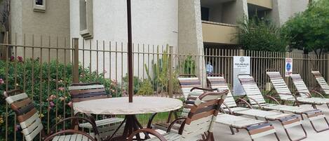 Un espace extérieur aménagé avec une table ronde et des chaises, un parasol en métal, et un coin détente au bord de la piscine avec des chaises longues.