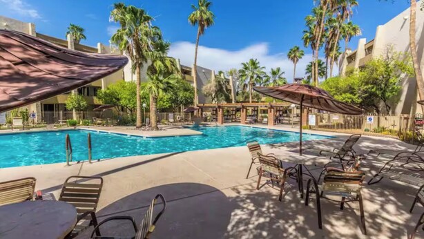 A pool area with lounge chairs and umbrellas, surrounded by palm trees and buildings.