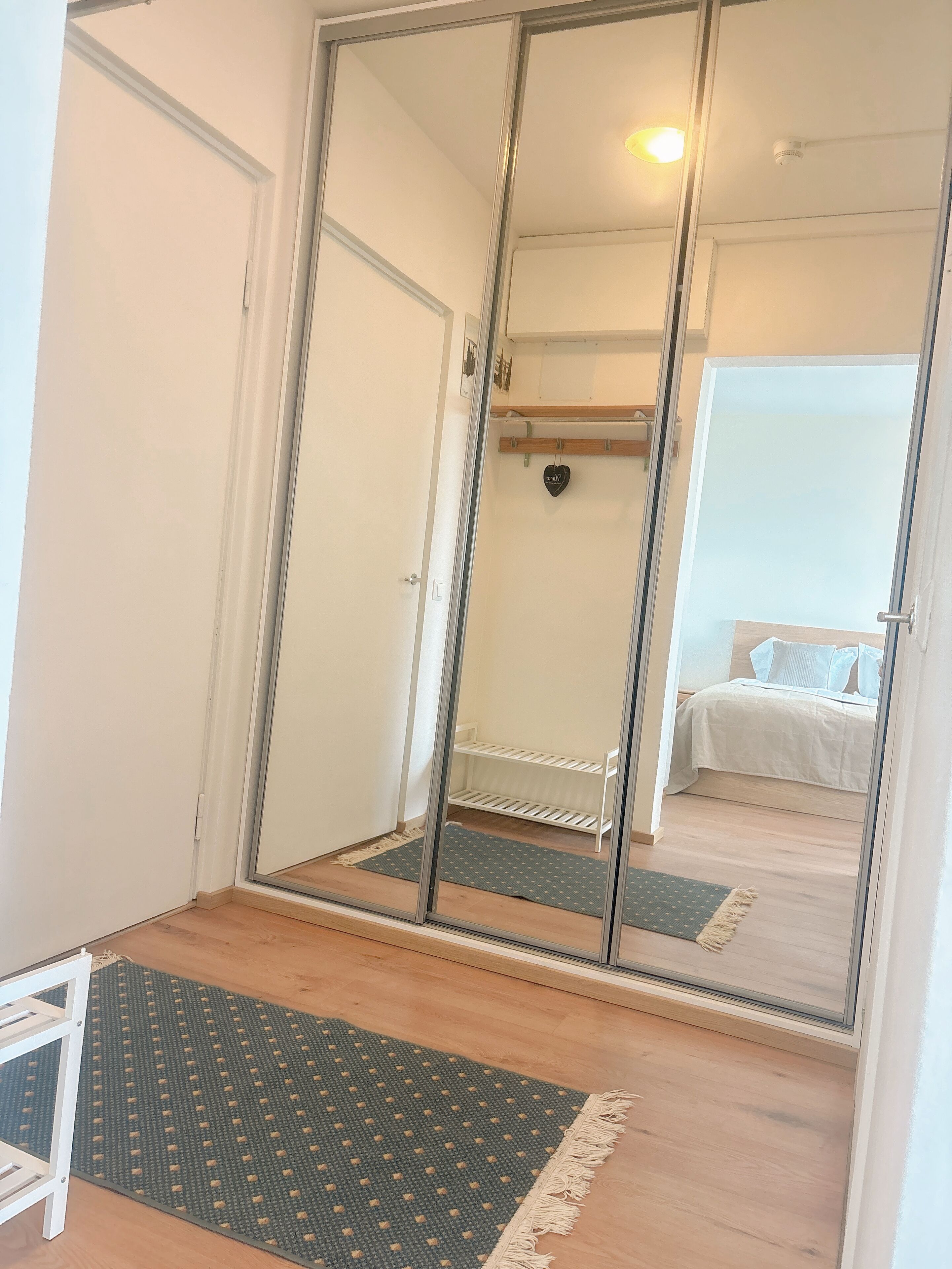 A room with a mirrored wardrobe, a bed with white bedding, a small shelf, and a mat on the floor.