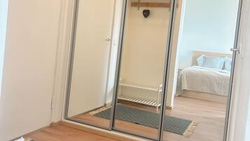 A room with a mirrored wardrobe, a bed with white bedding, a small shelf, and a mat on the floor.