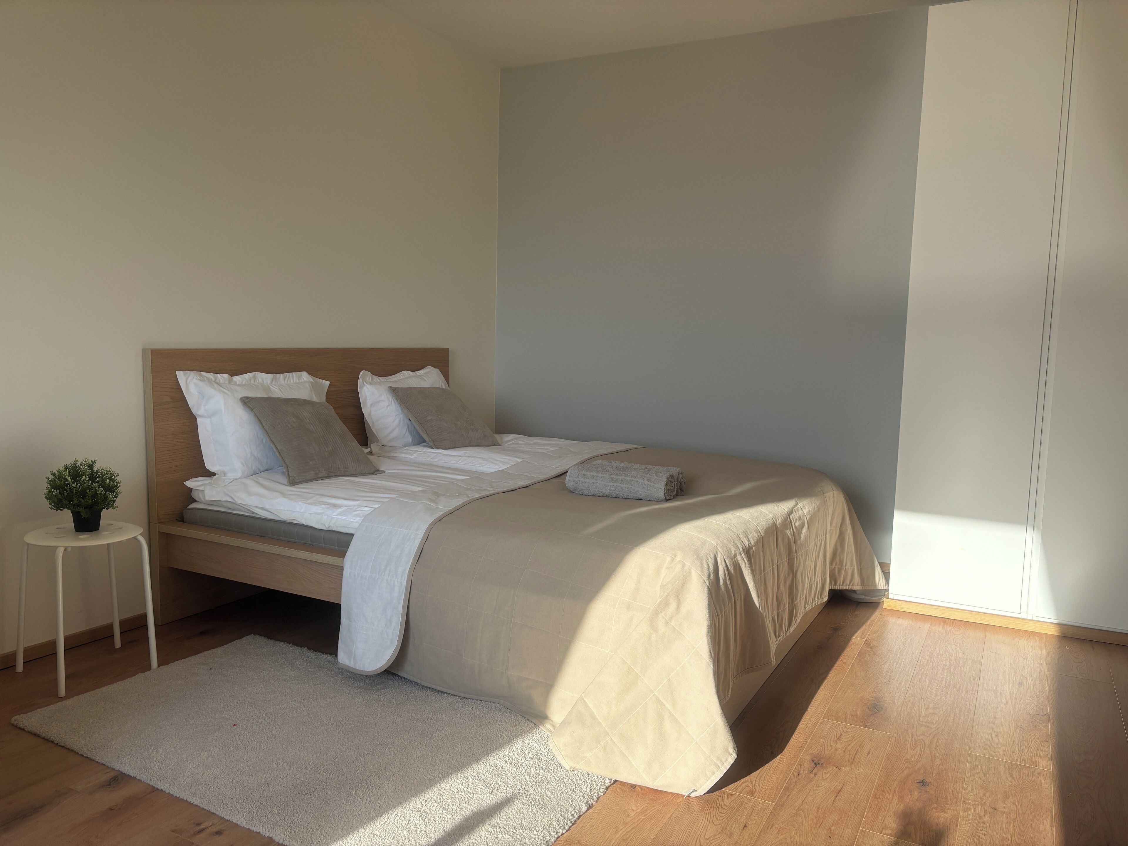 Ein ordentlich bezogenes Bett mit einem hölzernen Kopfteil, ein kleiner runder Tisch mit einer Pflanze und ein heller Teppich auf Holzdielen.