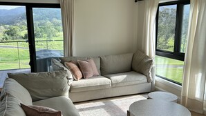 A modern living room with a sectional sofa, a round coffee table, and a ceiling fan. Large windows provide a view of a green landscape.