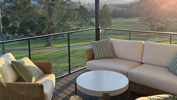 A covered outdoor seating area with wicker furniture and a round table, overlooking a scenic landscape with hills and trees.