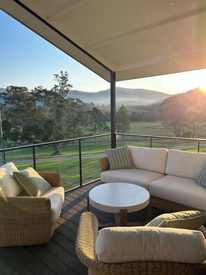 A covered outdoor seating area with wicker furniture and a round table, overlooking a scenic landscape with hills and trees.