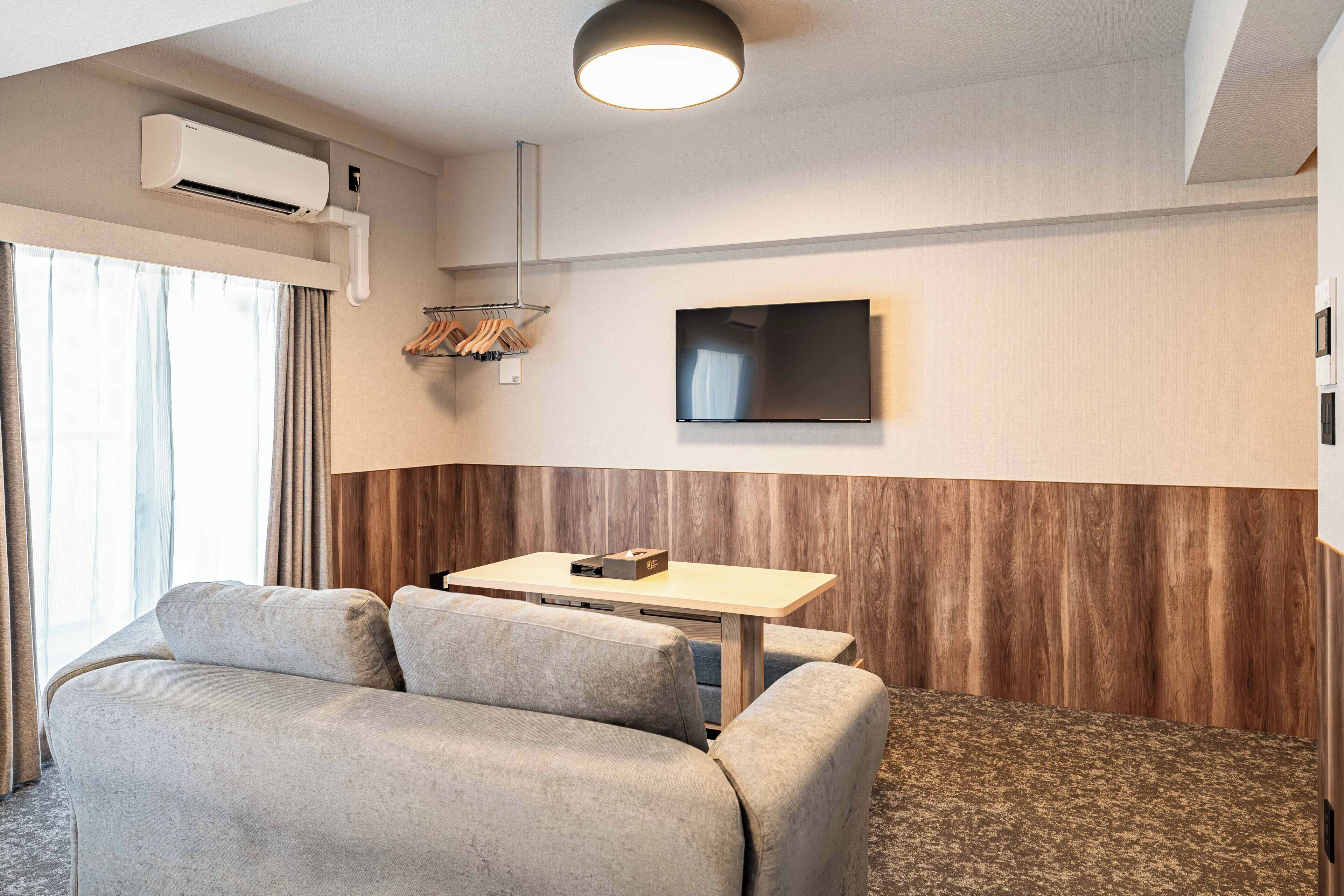 A modern hotel room with a sofa, a small table, a flat-screen TV, and an air conditioning unit.