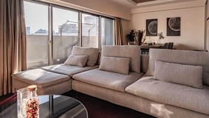 A modern living room with a sectional sofa, a glass coffee table, and a view of the city through large windows. - Apartment Hotel 11 Shinsaibashi (Osaka)