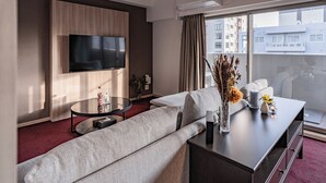 A modern hotel room with a large flat-screen TV mounted on the wall, a sofa, a coffee table, and a view of the city through large windows. - Apartment Hotel 11 Shinsaibashi (Osaka)