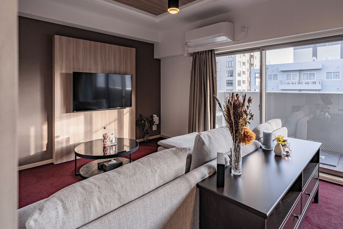 A modern hotel room with a large flat-screen TV mounted on the wall, a sofa, a coffee table, and a view of the city through large windows.
