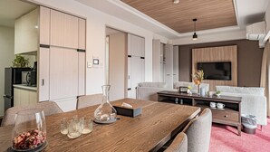 A modern living room with a wooden dining table, grey sofas, a TV mounted on the wall, and a kitchen area visible through an open door. - Apartment Hotel 11 Shinsaibashi (Osaka)