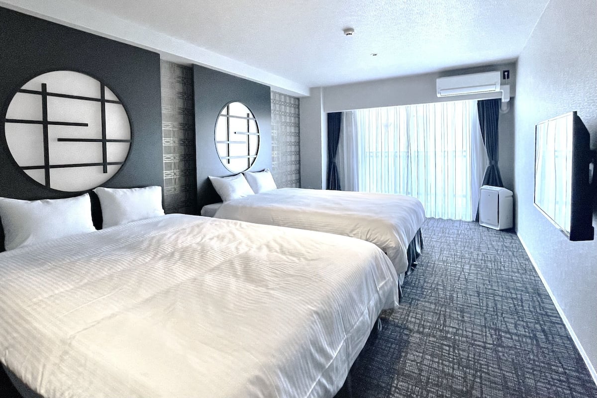 A hotel room with two beds, a large circular wall decoration, a large window with curtains, and an air conditioning unit.
