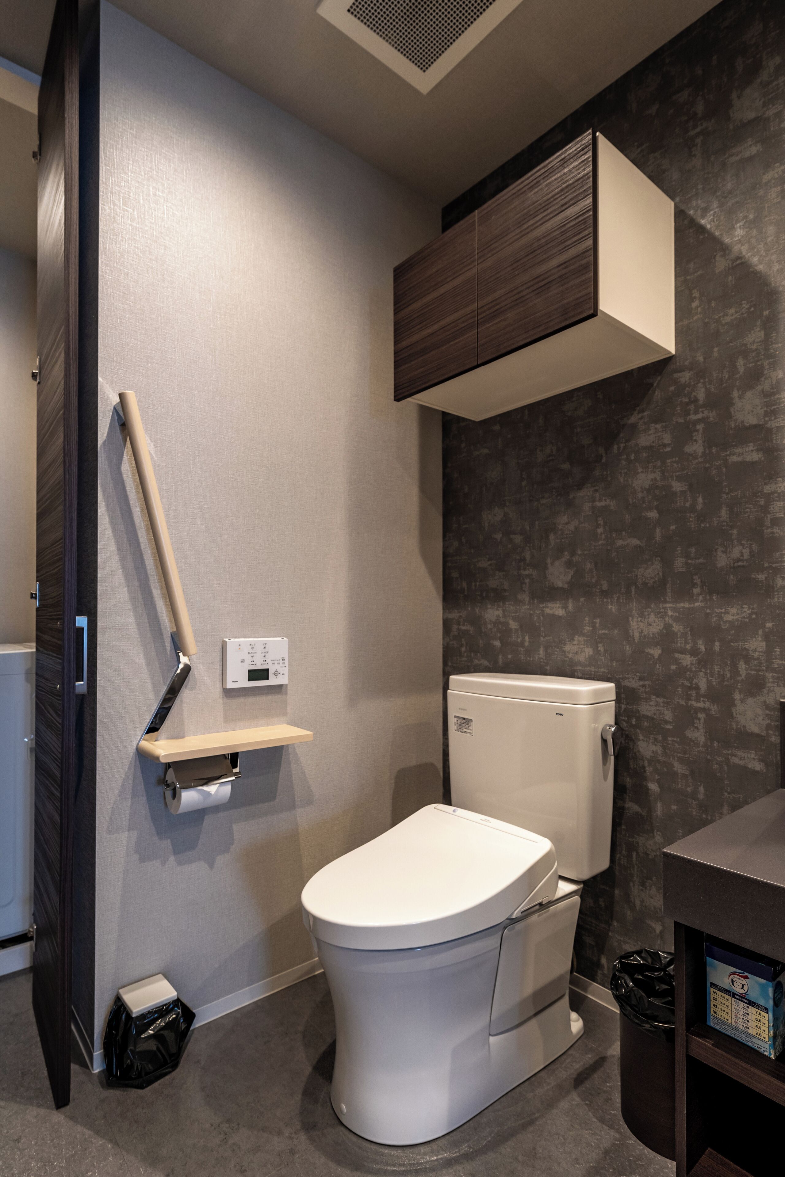 A modern bathroom with a toilet, a wall-mounted shelf, a hand dryer, and a trash bin.