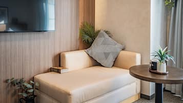 Quarto de hotel moderno com sofá bege, televisão de tela plana na parede, uma mesinha redonda com uma planta em vaso e outra planta ao lado do sofá.