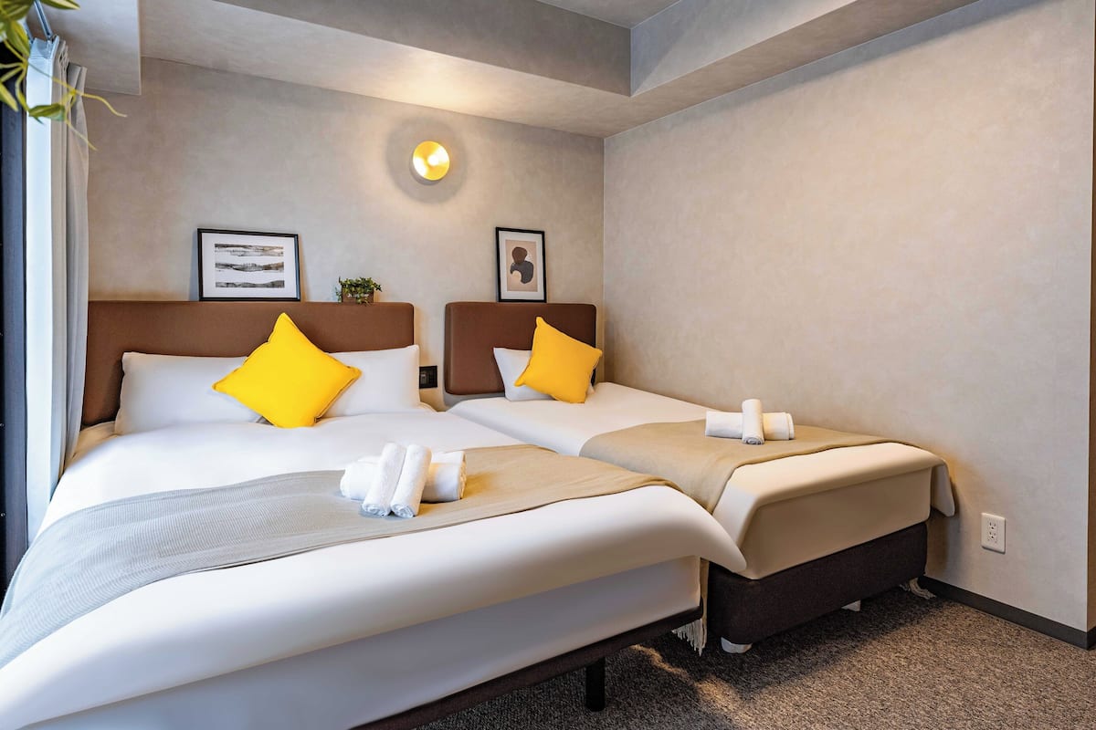Une chambre d’hôtel avec deux lits, chacun doté d’une tête de lit, de draps blancs et d’un oreiller jaune. Une petite plante est posée sur la tête de lit du lit situé à gauche, et un tableau encadré est accroché au mur au-dessus du lit situé à droite.