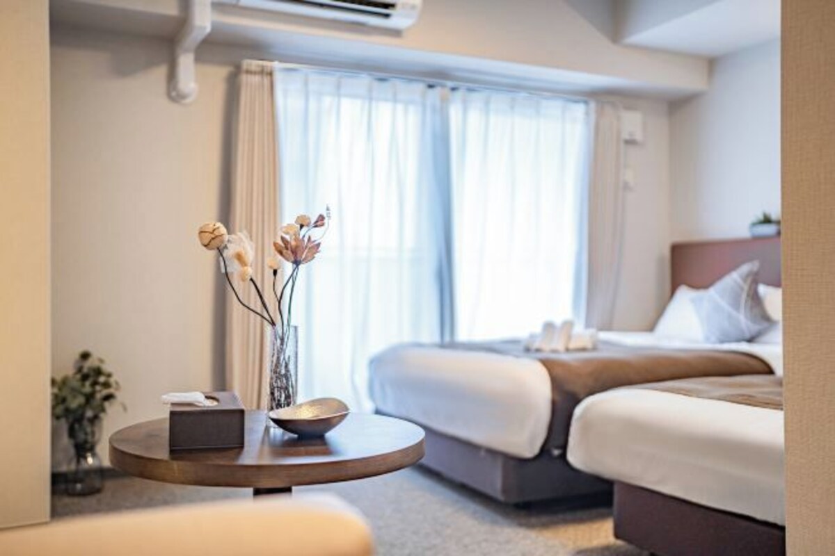 Une chambre d’hôtel avec un lit, une table ronde ornée d’un vase et d’un bol, et une fenêtre avec des rideaux.
