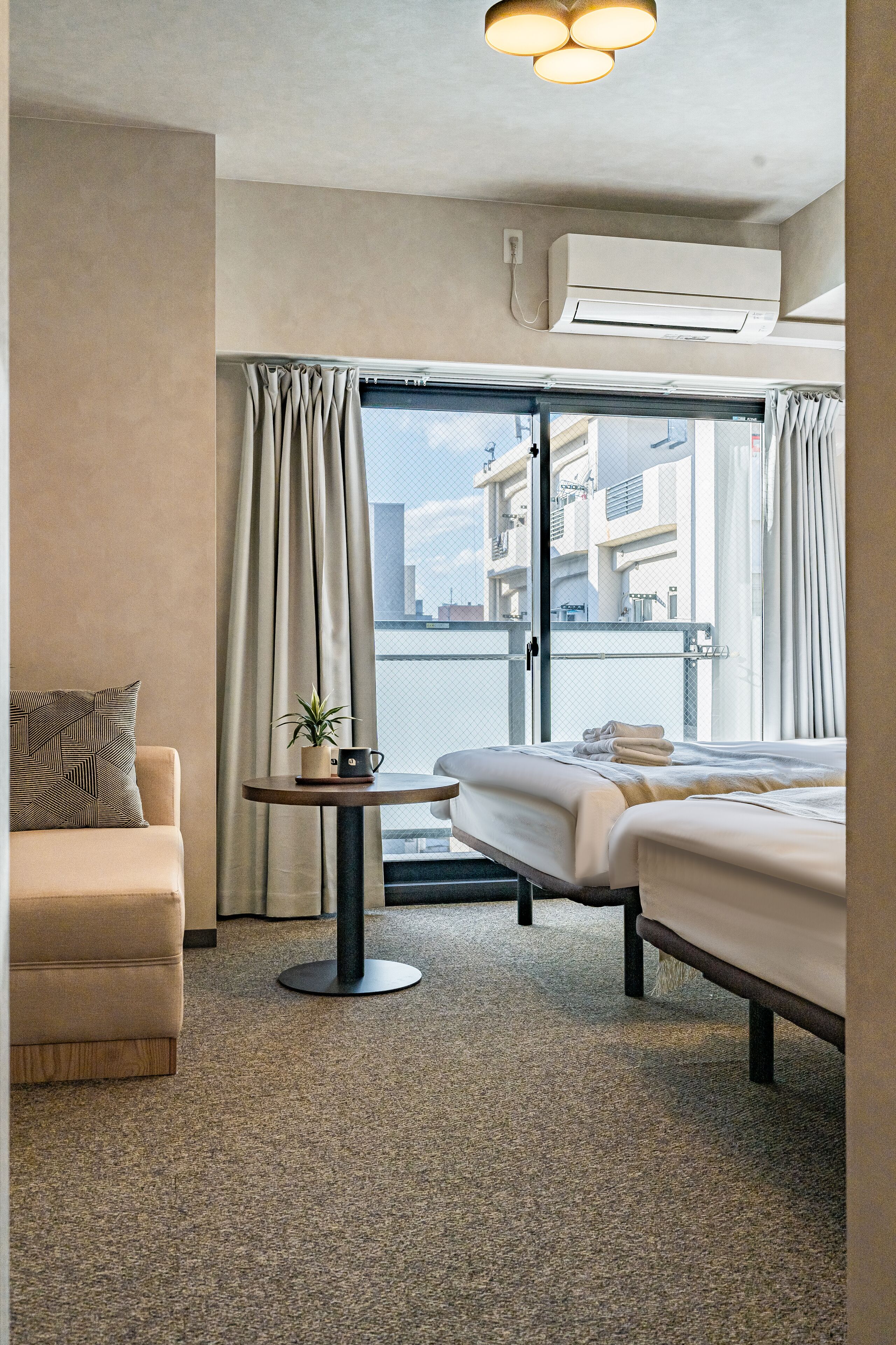A hotel room with a bed, a sofa, a small table with a plant and a cup, and a view of the city through large windows.