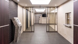 Un pasillo moderno de hotel con un ascensor de vidrio, una zona de recepción con un quiosco digital y un suelo de baldosas.