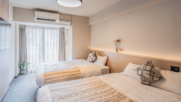 A hotel room with two beds, a ceiling light, an air conditioning unit, a window with curtains, and a small plant. - Apartment Hotel 11 Namba Minami III (Osaka)