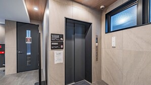 An elevator with a sign indicating a 10:00AM closing time, a security camera, and a window with a view. - Apartment Hotel 11 Namba Minami III (Osaka)