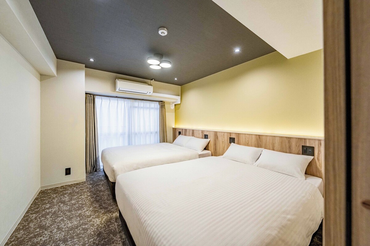 A hotel room with two beds, an air conditioning unit, a window with curtains, and a wooden headboard.