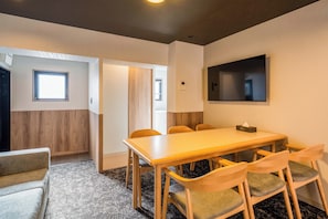 A conference room with a long table, six chairs, a flat-screen TV mounted on the wall, and a door leading to another room.