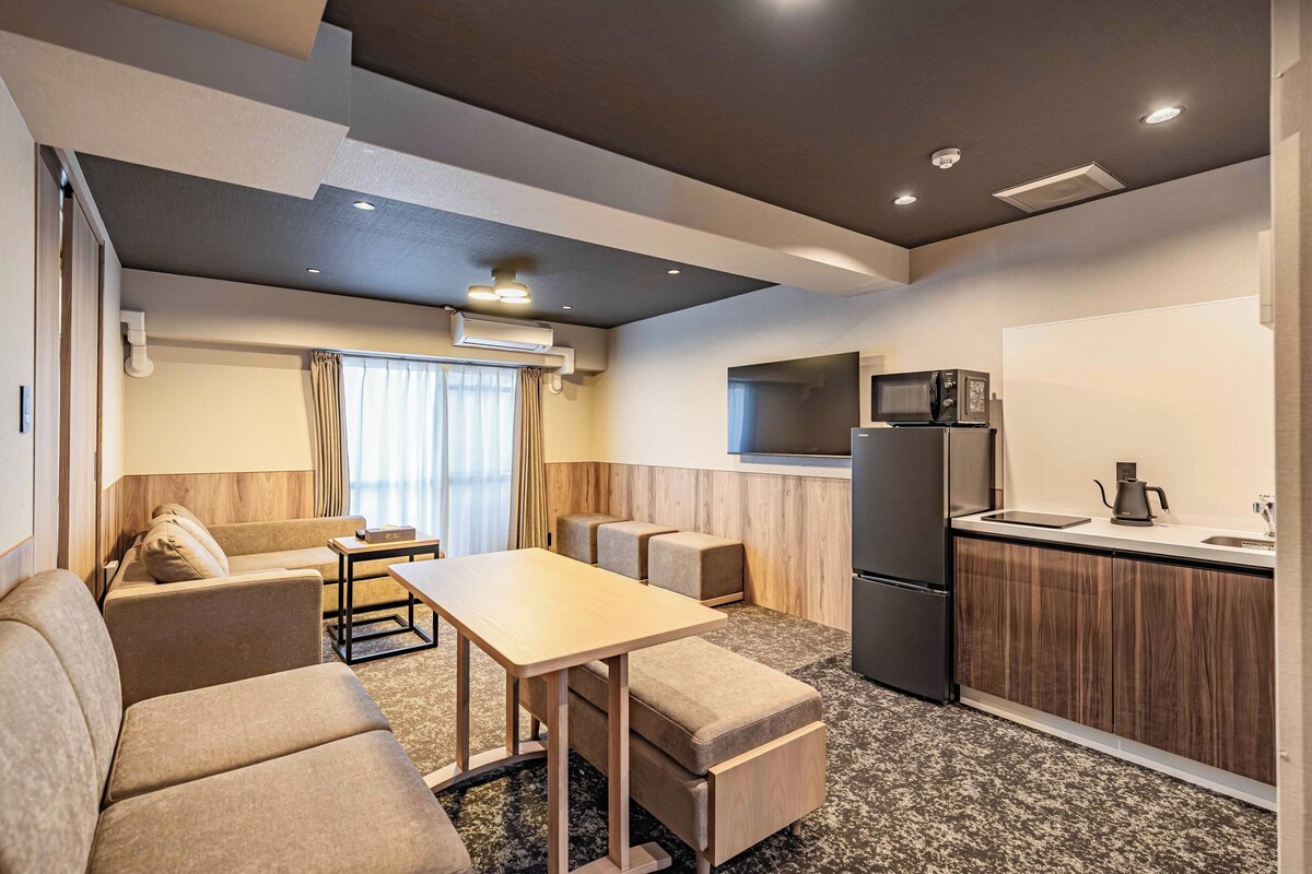 A modern hotel room with a sofa, a small dining table, a kitchen area with a microwave, and a refrigerator.